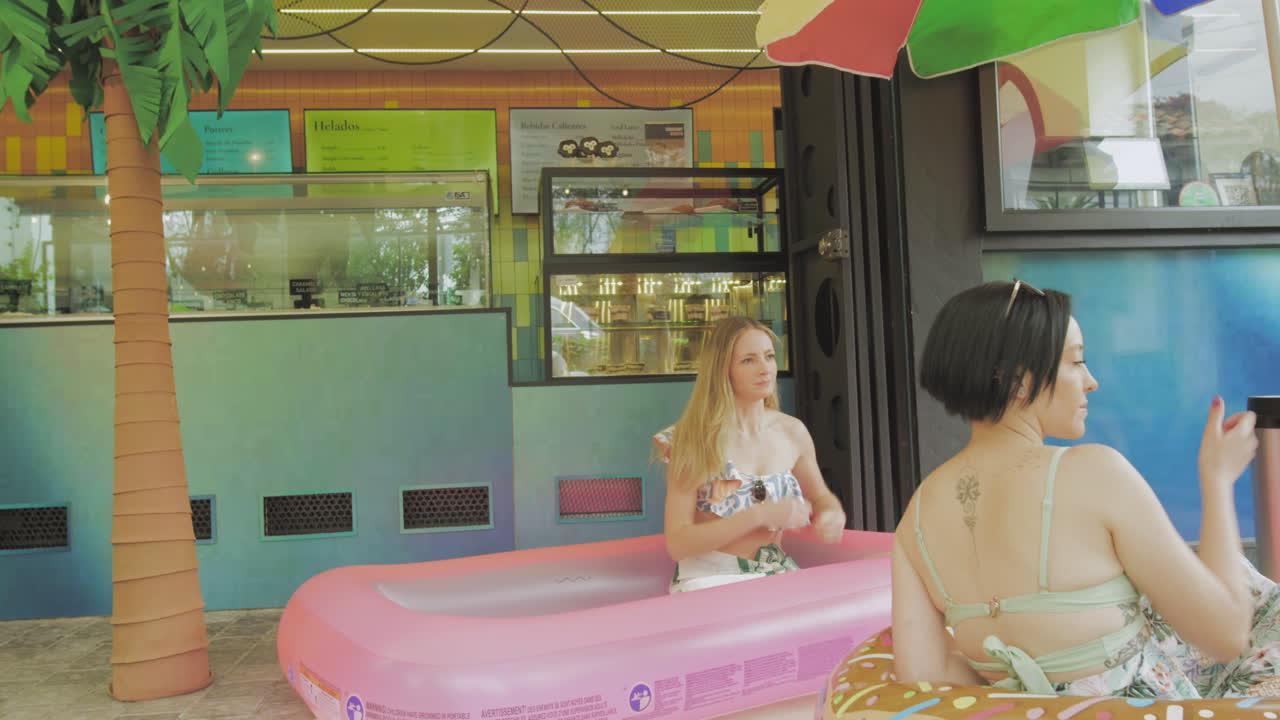 Two young women enjoy ice cream in a relaxed atmosphere next to an inflatable pool in front of a contemporary ice cream parlor.