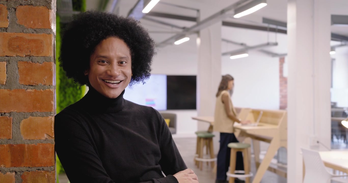 Homem afro-americano encostado em parede de tijolos lendo uma notificação de equipe diversa no celular, sorrindo no escritório.