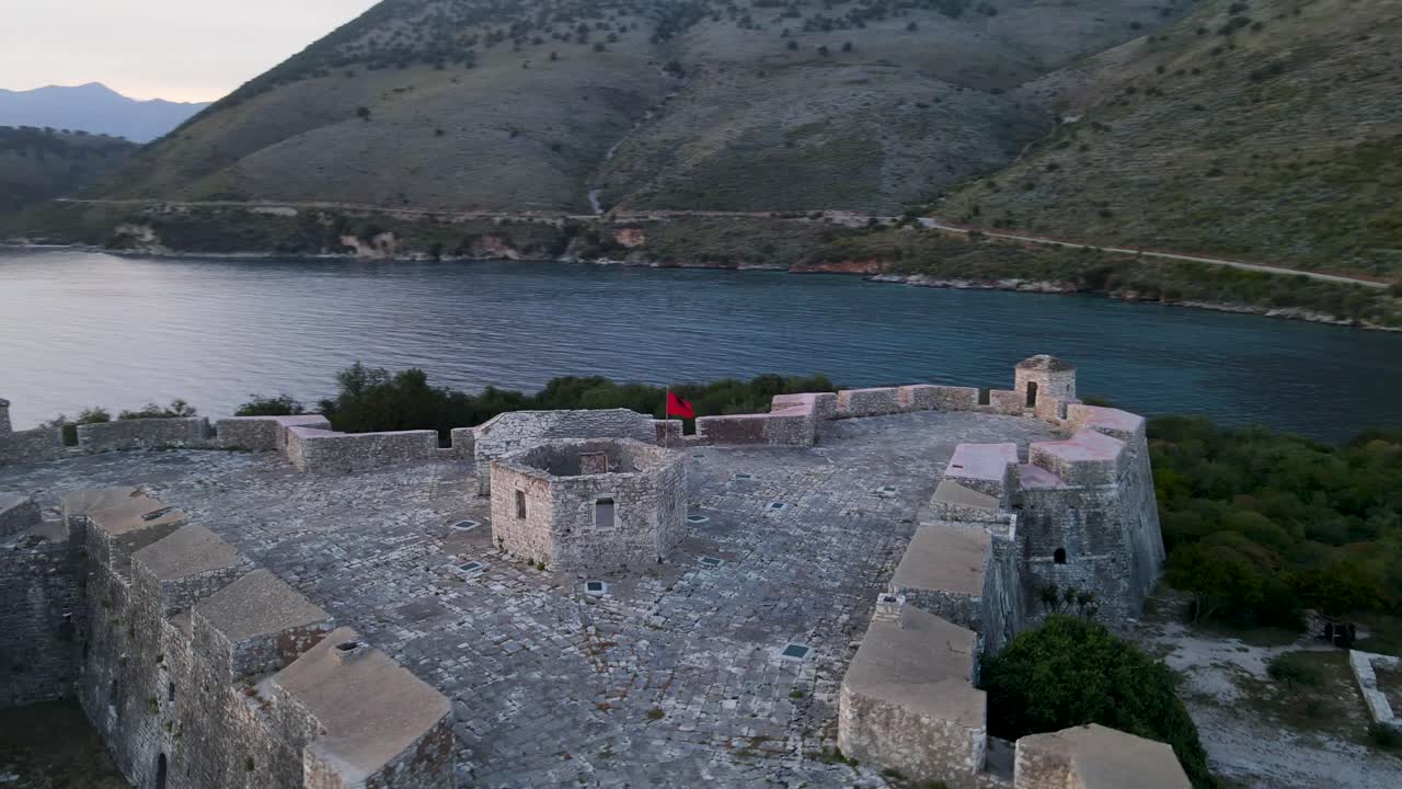 Vista aerea orbitante della bandiera albanese che sventola sulla cima del Castello di Porto Palermo nella affascinante Riviera albanese, mettendo in mostra l'orgoglio nazionale e il patrimonio storico
