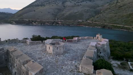 Aerial-orbit-view-of-fluttering-Albanian-flag-atop-Porto-Palermo-Castle-in-the-mesmerizing-Albanian-Riviera,-showcasing-national-pride-and-historical-heritage