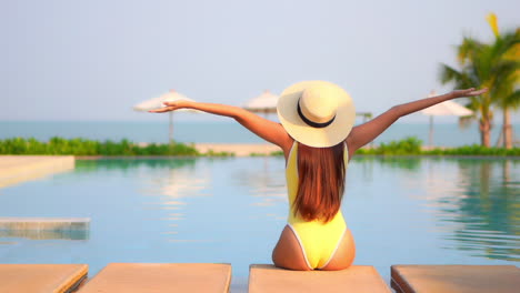 Vista-Trasera-De-Una-Mujer-Sexy-Sentada-Al-Lado-De-La-Piscina-En-Un-Hotel-Exótico-En-Bali-Con-Monokini-Amarillo-Y-Sombrero-Levantando-Las-Manos,-Ella-Levanta-Los-Brazos-Abiertos,-Palmeras-Borrosas-Y-Sombrillas-De-Playa