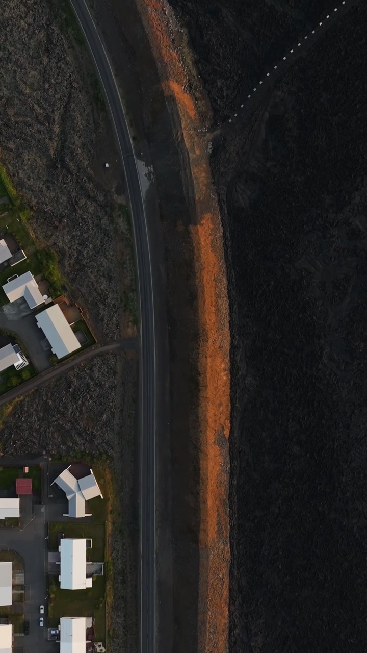 Vista verticale dall'alto con drone a Grindavik, Islanda, che mostra case e una strada curva protette da un vasto campo di lava nera da un lungo argine difensivo arancione.