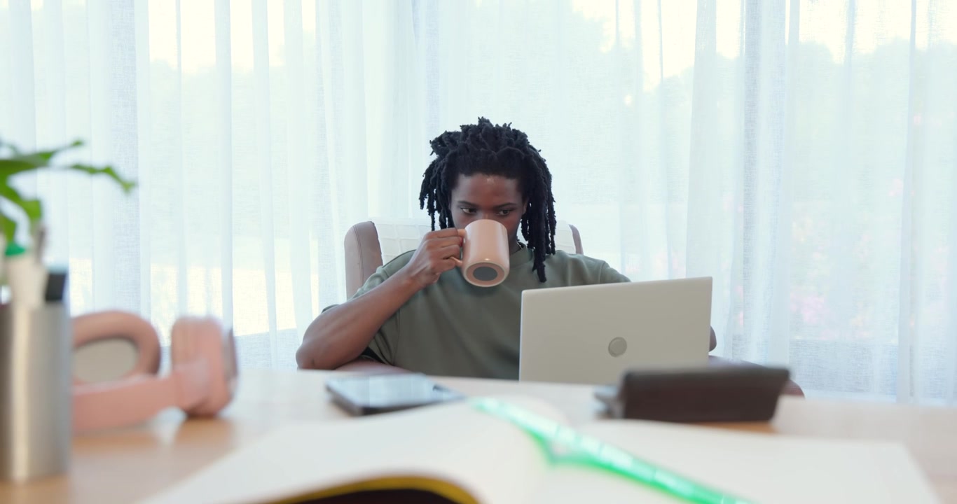 African American man in green tee sitting in armchair at home, sipping mug, reading laptop message