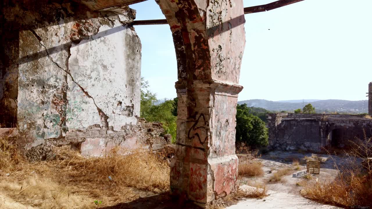 Ruinas de hacienda abandonada con paredes agrietadas y una antigua estructura de arco rodeadas de hierba seca y escombros bajo la luz brillante del sol en un paisaje rural.