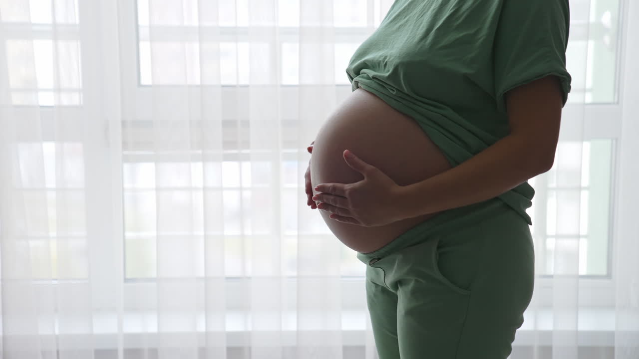 Pregnant woman standing near the window and relaxing at home. Maternity prenatal care and woman pregnancy concept