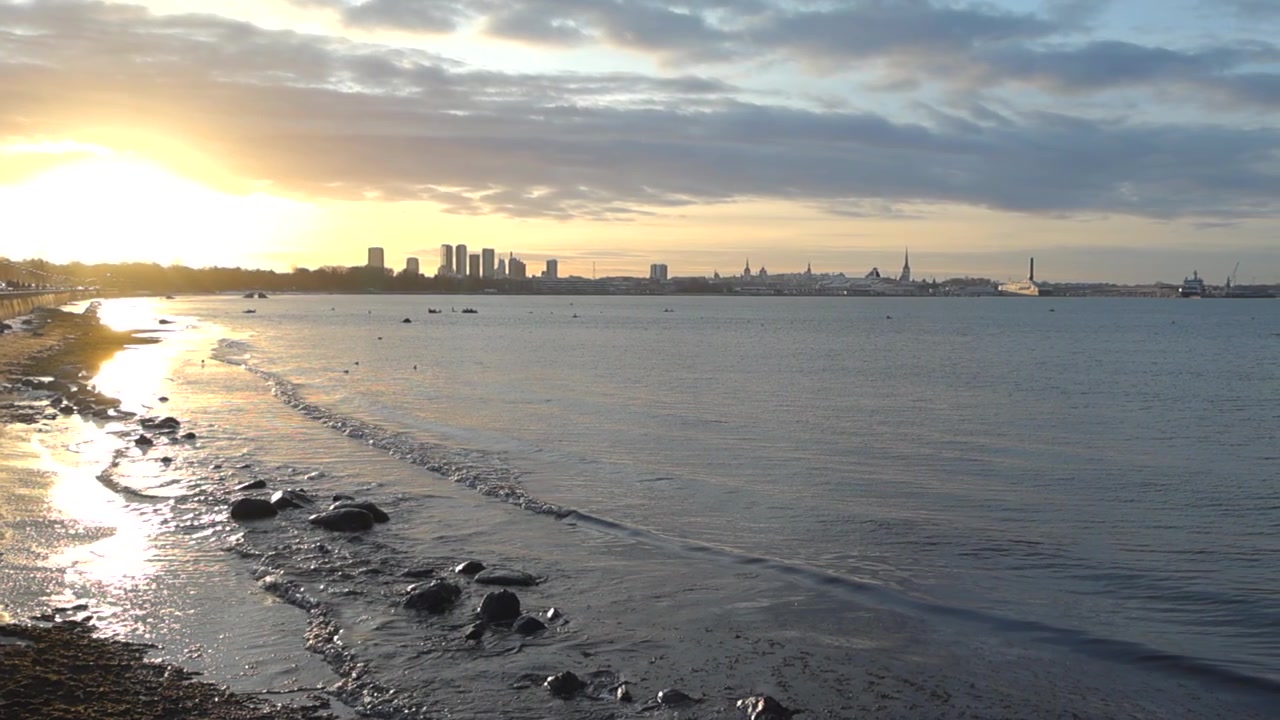 Ombak air pantai laut dengan riak air yang menuju ke pantai saat matahari terbenam yang memantul di air. Di latar belakang, siluet kota Tallinn terlihat dengan langit biru dan oranye keemasan