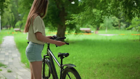 back view of woman with long hair approaches black bicycle parked on paved path, placing one glove on seat while wearing other, background features lush greenery and tall trees