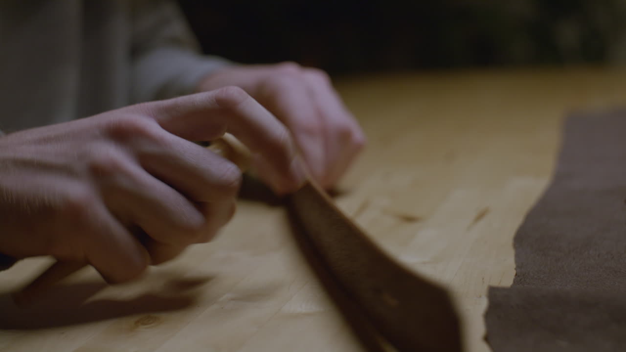 A close-up of hands stretching and shaping a brown leather strap on a wooden table, showcasing the detailed craftsmanship of leatherworking.