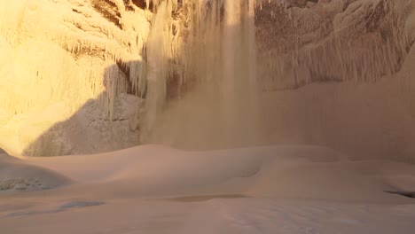 Tiro-Inclinado-Hacia-Abajo-De-La-Famosa-Cascada-De-Skogafoss-Cubierta-De-Nieve-Y-Hielo-Iluminado-Por-La-Puesta-De-Sol-Dorada