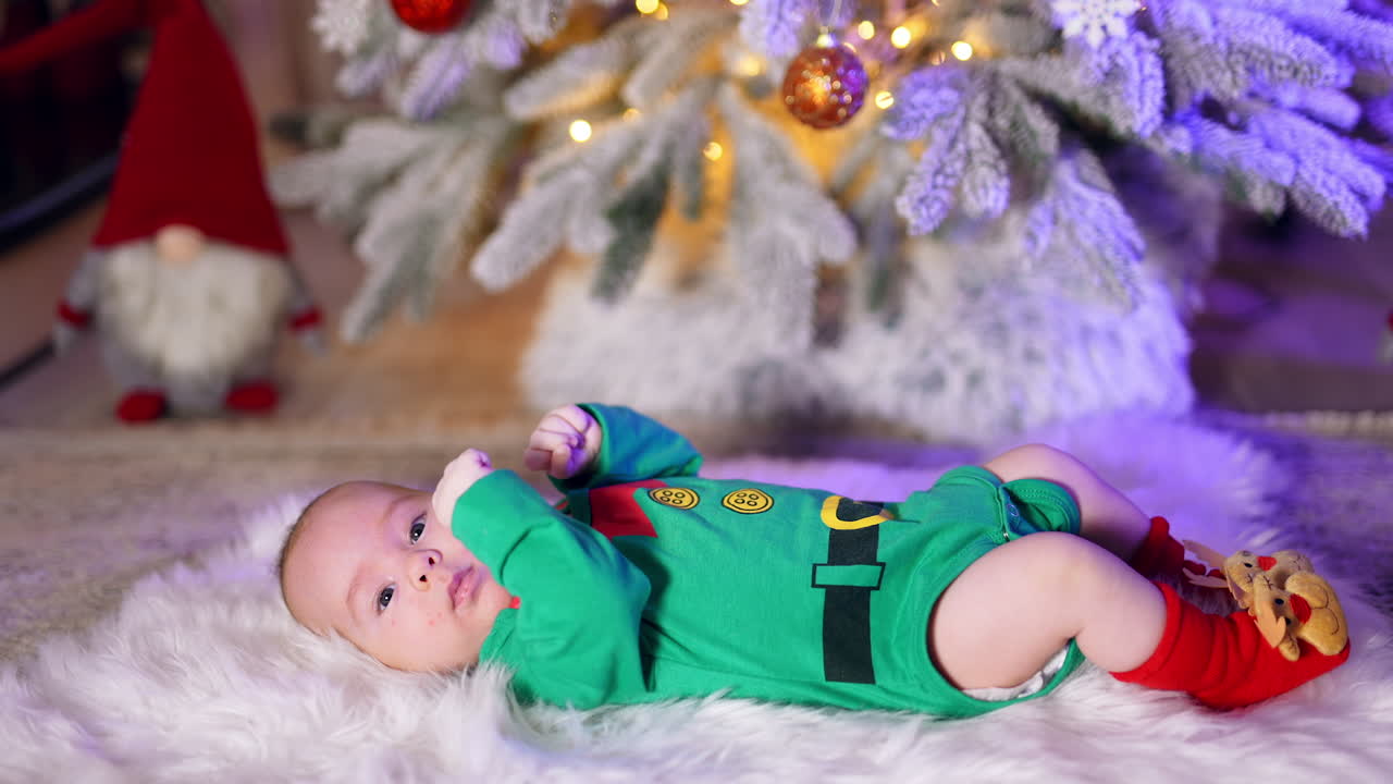 Adorable sweet kid in green costume lies peacefully on the plaid. Decorated Christmas tree at backdrop in blur.