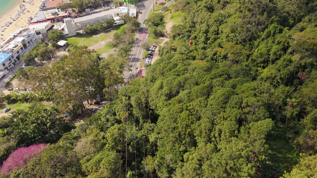 Downward angle drone approaches cable cars gliding above dense canopy of trees near ocean and coastline