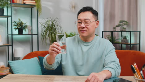 Un-Hombre-Asiático-Sediento-Se-Sienta-En-La-Mesa-De-Su-Casa-Sosteniendo-Un-Vaso-De-Agua-Natural-Y-Bebe-Sorbos-De-Agua-Filtrada.