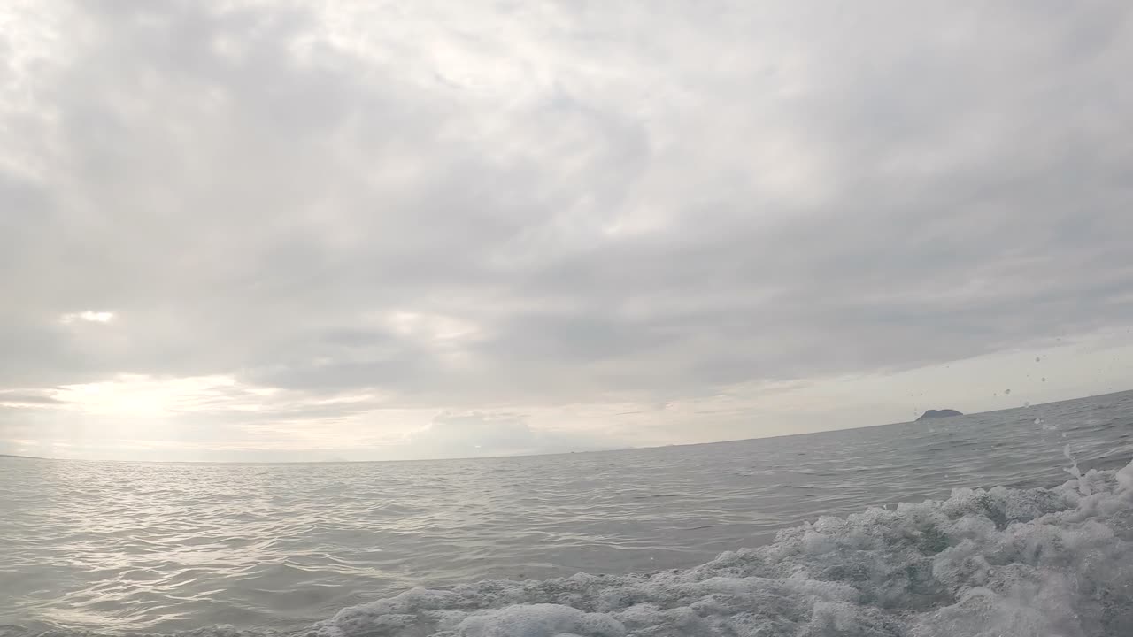 An island is seen on the back of an ocean of the Galapagos islands. There are waves formed by the panga ride. The sun is being covered by white clouds