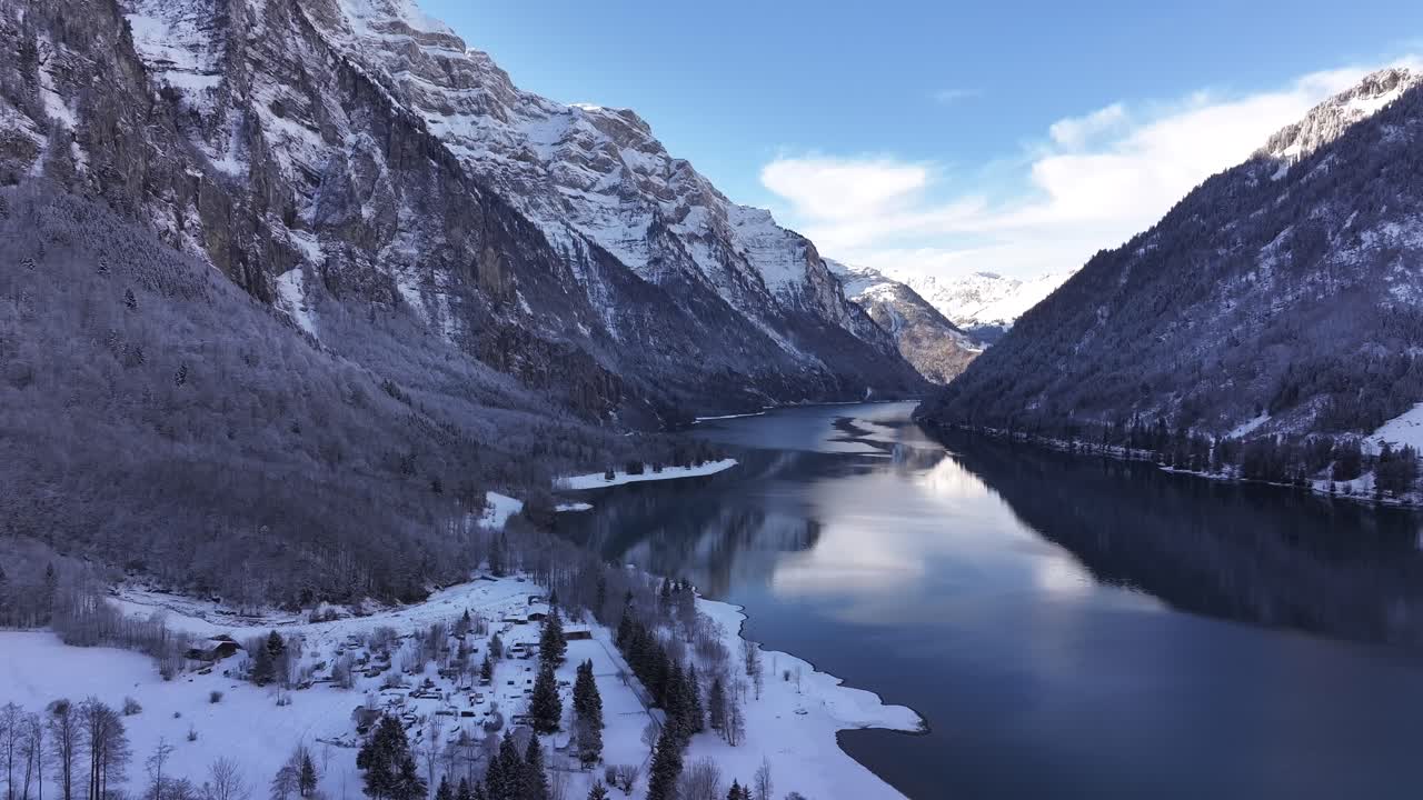 Dronelento Klöntalersee-järven yllä Sveitsin Alpeilla aurinkoisena päivänä, vuorilla on paljon lunta.