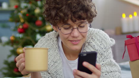 woman with short curly hair lying on the floor at home using smartphone in a room with christmas decorations