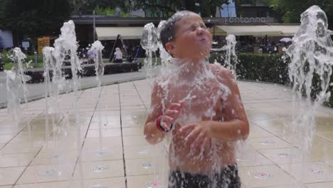 Slow-motion-shot-of-a-boy-running-through-the-fountains-of-a-city-fountain,-during-a-hot-summer-day