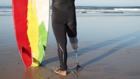 primer plano de un surfista masculino irreconocible en traje de neopreno con pierna artificial apoyado en la tabla de surf y parado frente al mar