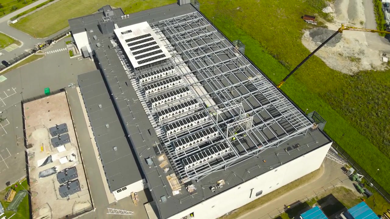 Industrielt byggeprojekt, der viser opførelsen af et stort datacenter, med en stålramme, der rejses på taget til kølesystemer, og tungt udstyr, der arbejder på stedet