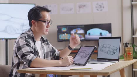 vista lateral de un hombre asiático pensando en un nuevo concepto de coche mientras trabaja en un boceto de diseño de coche en una tableta en el estudio con televisión y computadoras muestra modelo de coche eléctrico 3d
