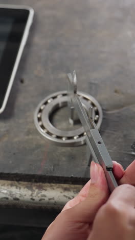 Overhead view of lab technician using vernier caliper to measure diameter of boris on workbench under dim light, recording measurements on tablet nearby. Industrial workspace with precision tools