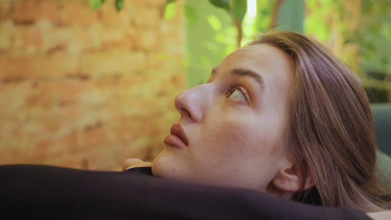Lady leans on cushion with head tilted upward, eyes gazing thoughtfully toward overhead greenery in cozy indoor space featuring warm brick wall and soft natural light through surrounding plants