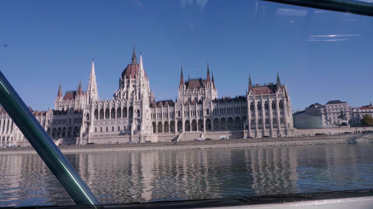 Vista da un battello fluviale mentre il Parlamento di Budapest scorre all'esterno. Ringhiere e riflessi delle finestre visibili
