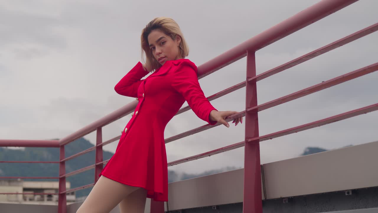 In Port of Spain, Trinidad, a young girl of Hispanic descent stands on a rooftop, dressed in red, with cloudy skies in the distance low angle