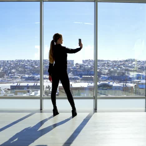 Young woman taking selfie photo in the office. Beautiful girl standing by the large panoramic windows and taking photo of herself on a phone. Rear view