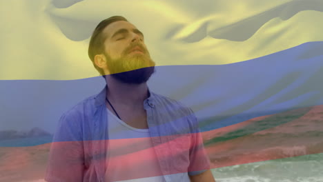 video compuesto de agitar la bandera de colombia sobre el hombre caucásico con los ojos cerrados de pie en la playa