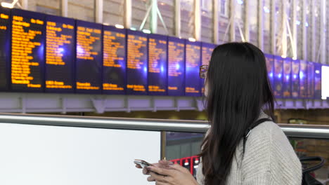 mujer joven en su teléfono revisa el tablero de salida de la estación de tren antes de salir para tomar su tren