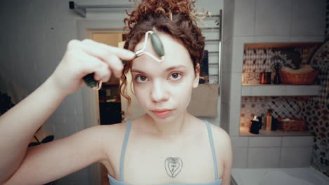 una joven se da un masaje facial con un rodillo de jade en el baño.
