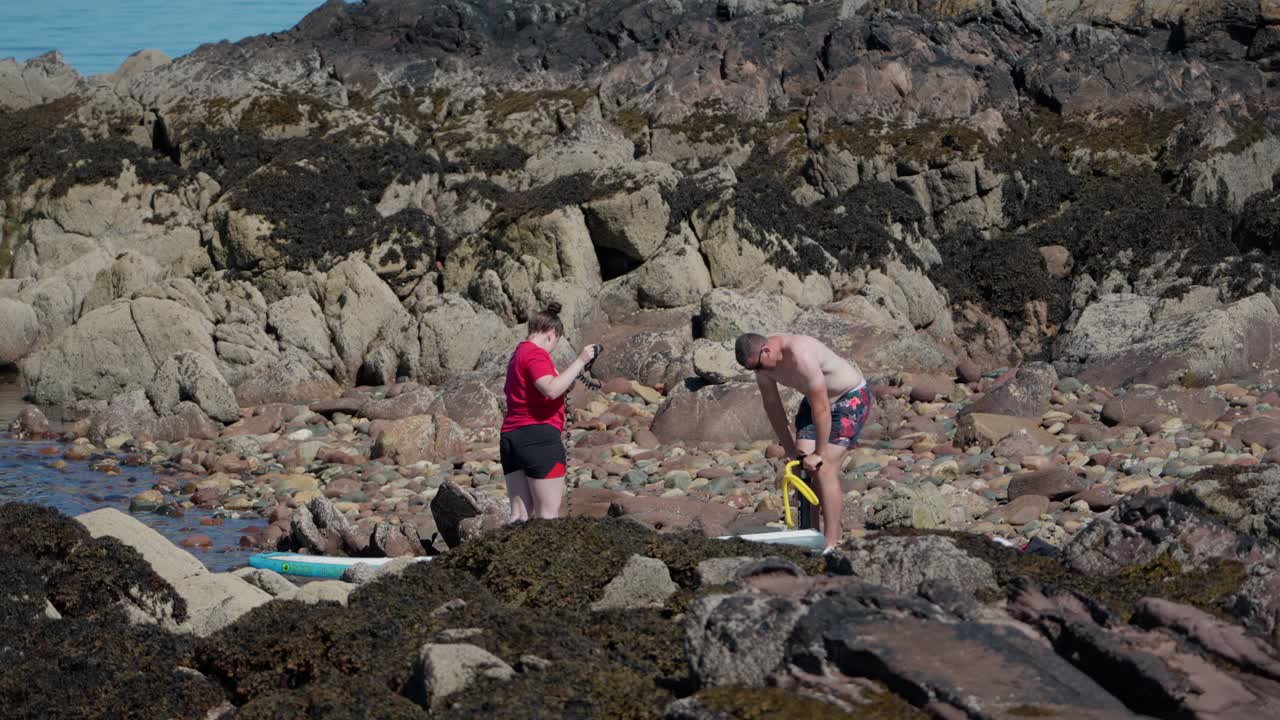 Young Man and his girlfriend on a rocky shoreline inflates his paddle-board next to rugged seaside terrain, ideal for active travel and adventure themes