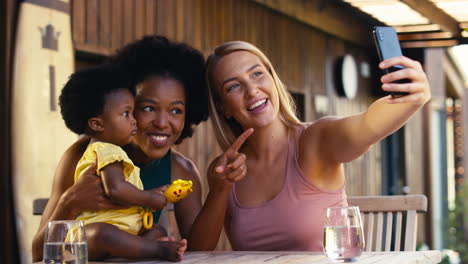 Same-Sex-Family-With-Two-Mums-Taking-Selfie-With-Young-Daughter-Outdoors-At-Home