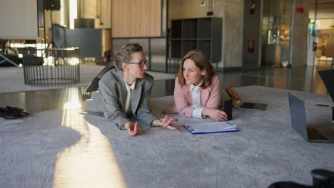 Middle-aged-blondes-in-business-clothes-lie-on-the-floor-on-a-gray-soft-carpet-and-communicate-during-a-break-at-work.-In-the-office