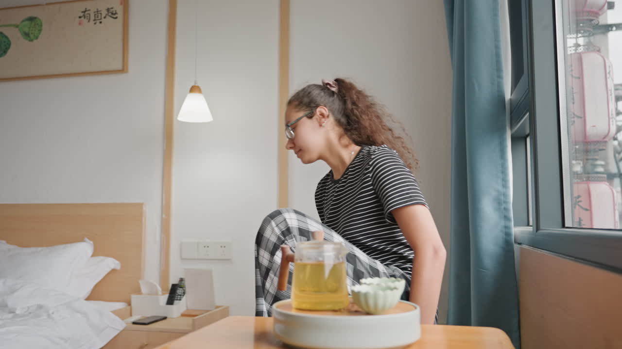 Mattinata rilassata con tè e luce solare delicata. Prepara il tè accanto al letto mentre la luce del sole filtra attraverso le tende. L'ospite si rilassa e prepara il tè durante una mattinata tranquilla con la luce del sole che entra