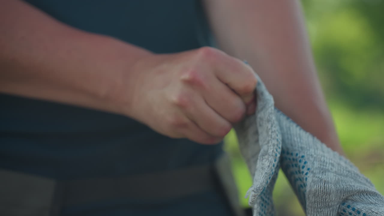 close up fair skinned man putting on glove from left hand, fingers adjusting fit, soft green background blur, sunlight highlighting skin texture, preparation for outdoor work