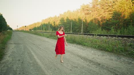 woman in red floral dress walking casually along countryside dirt road near railway track, surrounded by tall grass and pine trees under soft evening light in peaceful rural setting