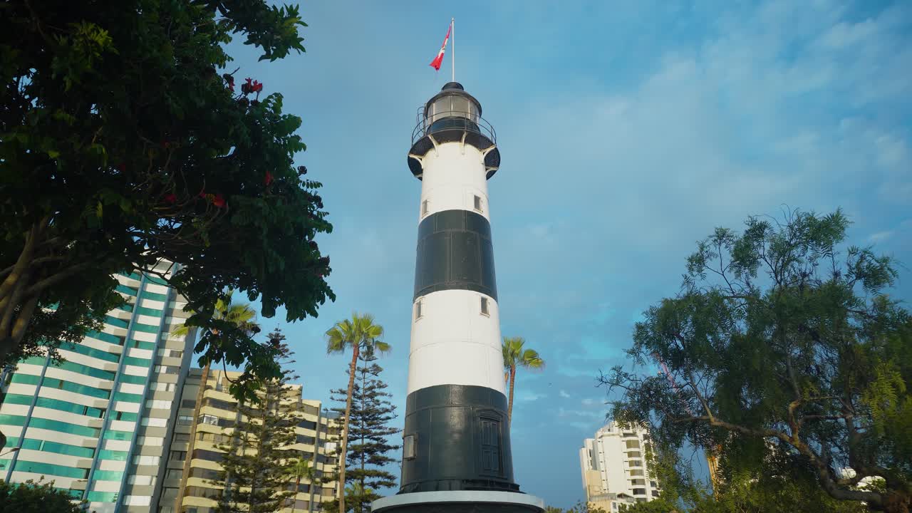 Una ripresa statica 4K di un faro colorato in bianco e nero con la bandiera peruviana in cima, circondato da alberi in un parco pubblico con condomini dietro. Situato nel quartiere Miraflores di Lima, Perù.
