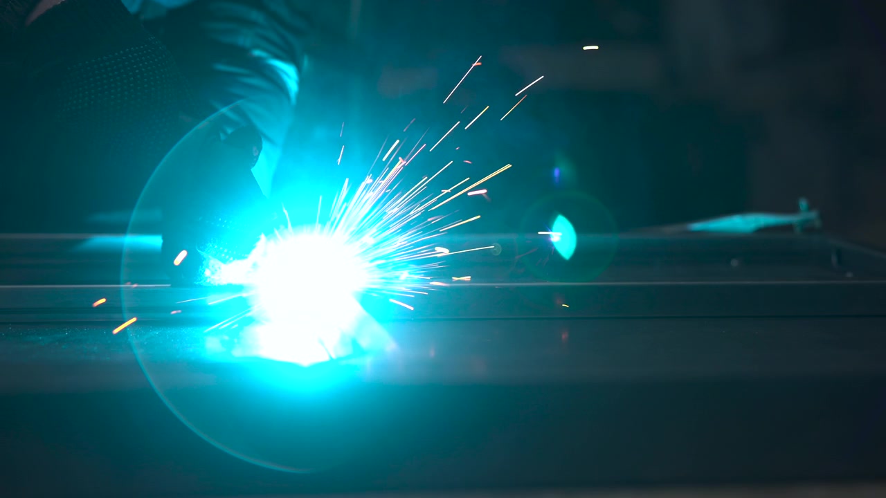 Worker silhouette leans toward metal surface during bright welding, with blue white arc, orange sparks, reflective steel, deep shadows, and low angle industrial workshop setting creating focused