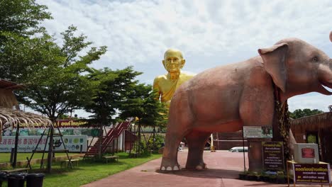 Panning-shot-showing-bright-golden-monk-Luang-Pu-Thuat-statue-in-the-background-and-a-giant-elephant-sculpture-in-the-foreground-at-the-Buddhist-Park-in-Phuttha-Utthayan-Maharat-Ayutthaya-Thailand