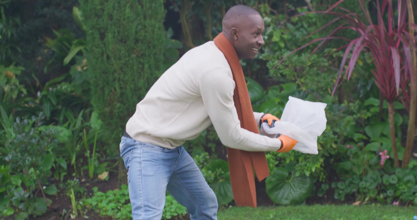 Spotting litter, African American man in orange gloves picking bottles into white bag to clean yard