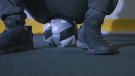 partial view of person dressed in black, seated on soccer ball with black sneakers on turf, yellow marking on the metallic fence in background