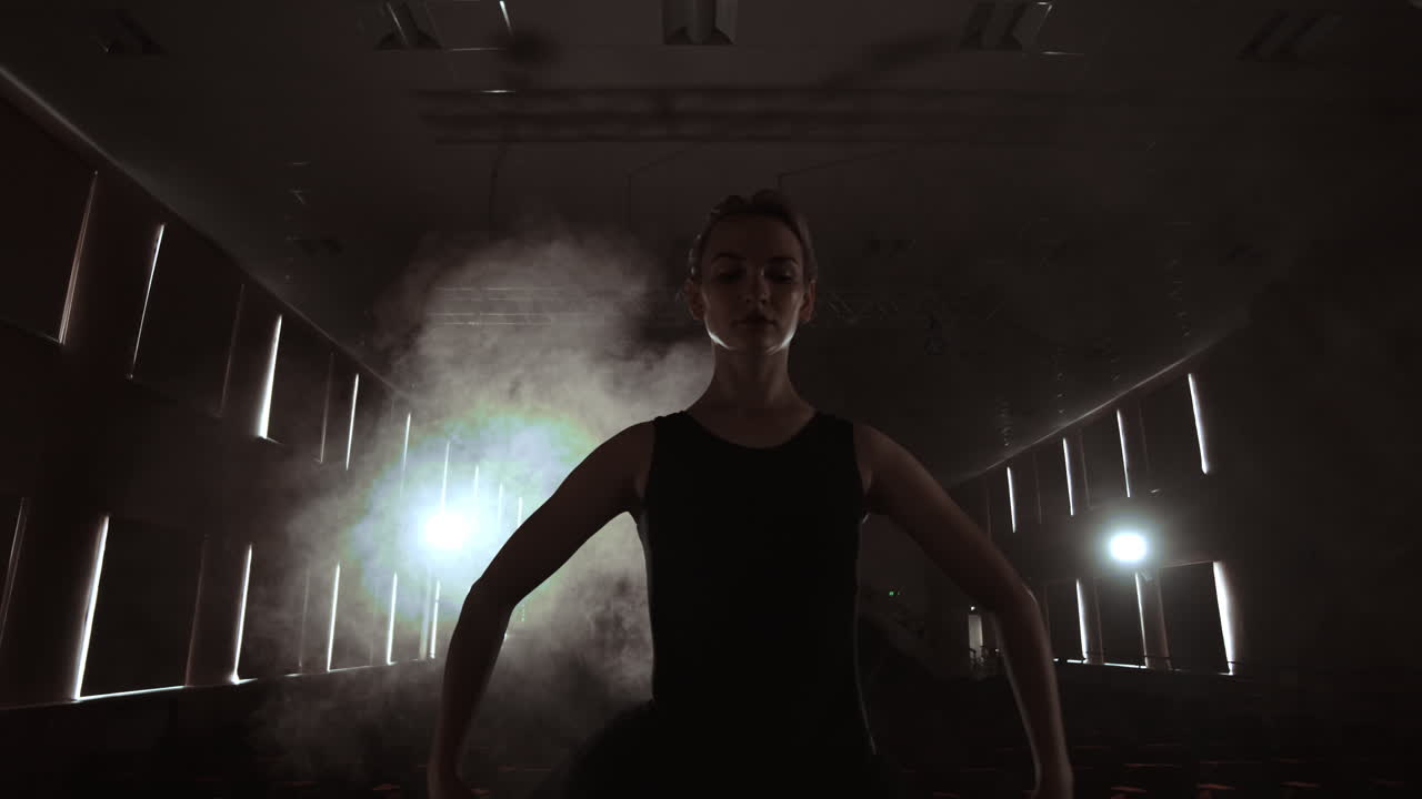 SLOW MOTION: prima ballerina prova un assolo in piedi sul palco del teatro, nel fumo, in abito nero e sotto i riflettori