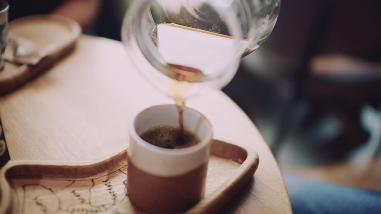 Nærbillede af friskbrygget kaffe, der hældes fra en glaskande ned i en keramikkop på en træbakke.