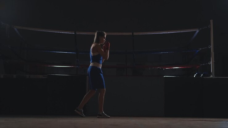 Una bellissima pugile professionista sferra pugni facendo shadow boxing in una sala buia di una palestra, al rallentatore, con abiti blu e bende rosse ai polsi. Oggetti che volteggiano. Ripresa con steadicam.