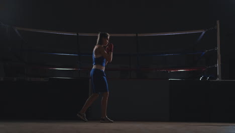 Professional-beautiful-female-boxer-otbryvatyvat-blows-conducting-a-fight-with-a-shadow-in-a-dark-hall-of-the-hall-in-slow-motion-in-blue-clothes-and-red-bandages-on-her-wrists.-flying-around-the-object.-steadicam-shot