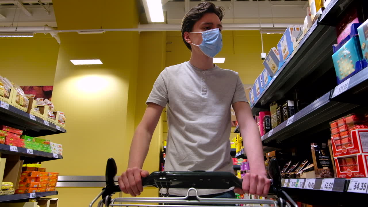 CHISINAU, MOLDOVA - JUNE 5, 2020: Young man with a protective medical mask walking in a supermarket near the shelves, taking some sweets and putting in the cart. Corona Virus idea