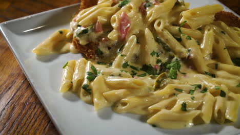 tabletop view of creamy penne pasta over fried chicken cutlet on square restaurant dish, close up slider 4k