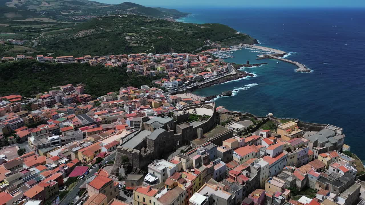 Scenic aerial view of coastal Mediterranean town with old castle, colorful rooftops, and harbor beside deep blue sea under clear daylight sky surrounded by green hills