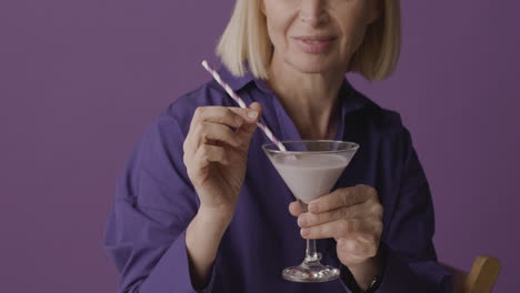 blonde mature woman with purple shirt posing holding a cocktail and smiling at camera on purple background 1